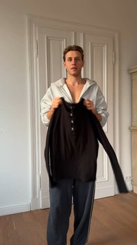 Musta pitkähihainen paita napillinen etukappale, jota pitelee harmaaseen huppariin pukeutunut henkilö neutraalia taustaa vasten.