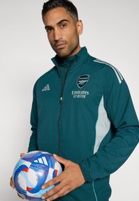 Tilausturkoosinvärinen vetoketjullinen takki harmailla yksityiskohdilla ja Arsenal-logolla; pitää kädessään sinivalkopunaista kuvioitua jalkapalloa. Sileä pinta, urheilullinen istuvuus.