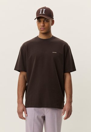 Mężczyzna ubrany w ciemnobrązowy T-shirt z napisem "LES DEUX" oraz pasującą brązową czapkę z białym "II", połączone z jasnoszarymi spodniami, na jednolitym tle.