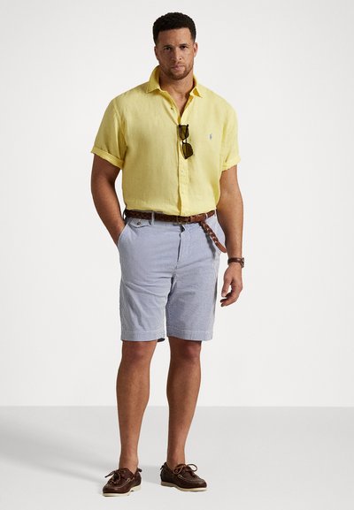 Chemise jaune à manches courtes en lin, shorts rayés bleu et blanc, ceinture en cuir marron, chaussures bateau marron et lunettes de soleil suspendues au col.
