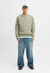 Lichtgroene sweatshirt met ronde hals, gecombineerd met lichtblauwe oversized jeans. Bruine schoenen en een witte pet met groene accenten.