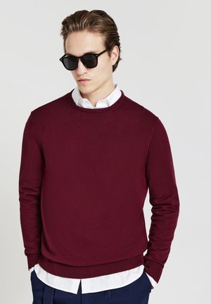 Weinroter Rundhalsausschnitt-Pullover mit langen Ärmeln, strukturiertem Stoff und geripptem Saum, getragen über einem weißen Hemd, kombiniert mit dunklen Sonnenbrillen.