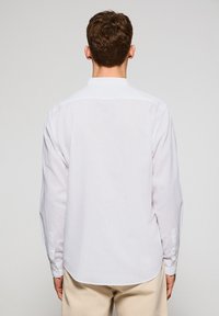 Vit långärmad skjorta med nedknäppt krage, tillverkad av ett lätt material. Den har en rundad nederkant och manschettknappar, visad bakifrån.