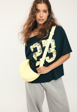 Junge Frau mit langen, welligen Haaren, die ein schwarzes, übergroßes T-Shirt mit der Aufschrift "23", graue Hosen und eine hellgelbe Umhängetasche trägt.