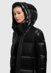 Schwarze Pufferjacke mit glänzender Oberfläche, großer Kapuze, hohem Kragen und kontrastierenden Akzenten. Mit Reißverschluss versehene Taschen an den Seiten.
