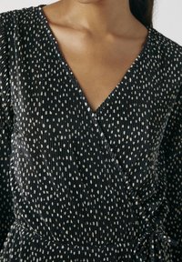 Femme portant une robe noire plissée enroulée avec des pois irréguliers blancs et un décolleté en V, nouée à la taille.