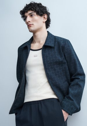 Νεαρός με σγουρά μαλλιά, φοράει σκούρο σακάκι με σχέδια πάνω από λευκό αμάνικο μπλουζάκι και σκούρο παντελόνι, στέκεται μπροστά σε ανοιχτό γκρι φόντο.