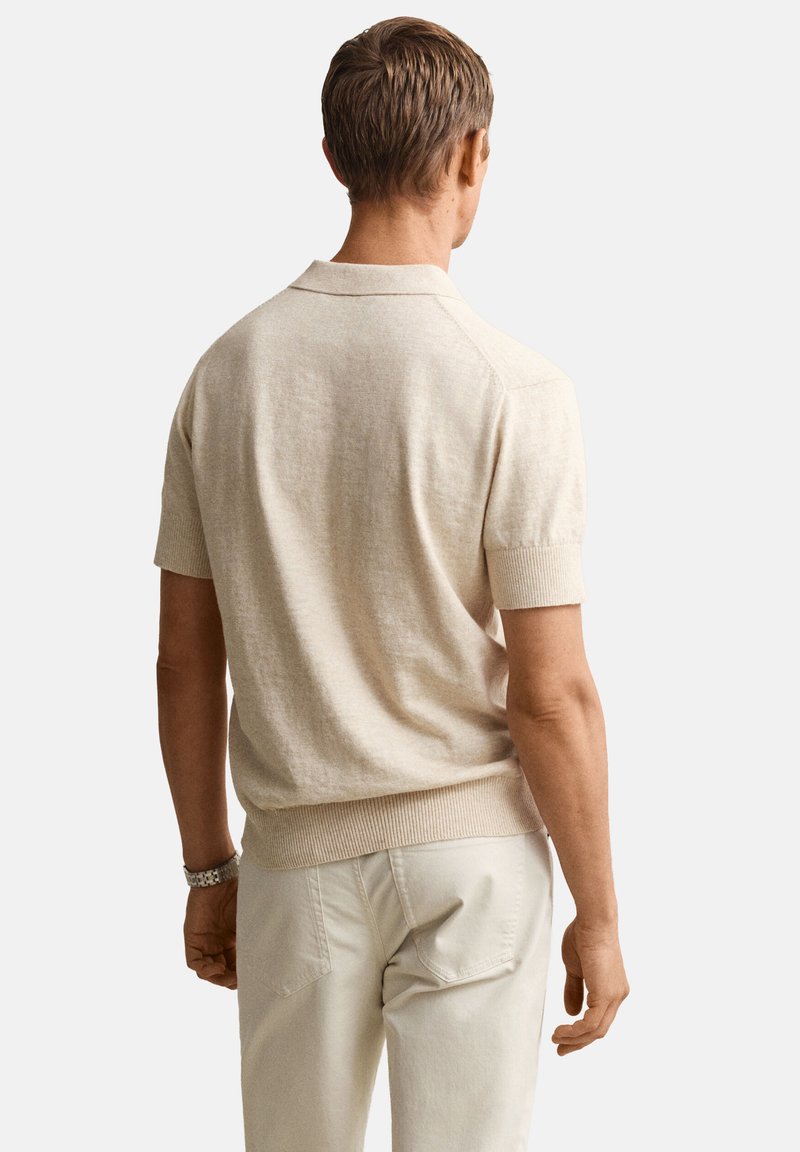 Uomo con polo beige a maniche corte in maglia e pantaloni chiari, in piedi di spalle contro uno sfondo semplice.