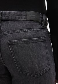 Fekete farmernadrág sima textúrával, téglalap alakú hátsó zsebbel és márkás bőrpánttal a derékon.