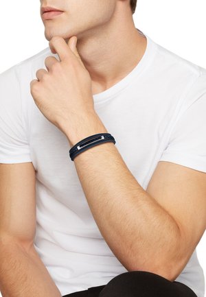 La pulsera azul marino presenta una textura trenzada y un acento metálico, llevada en una muñeca sobre una camiseta blanca lisa.