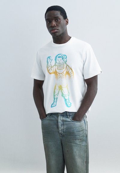 Giovane uomo con una T-shirt bianca raffigurante un astronauta dai colori arcobaleno, in piedi con le mani in tasca davanti a uno sfondo semplice.