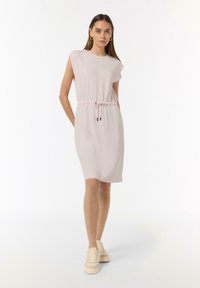 Helles rosa, knielanges Kleid mit kurzen Ärmeln, Bindeband in der Taille und glattem Stoff. Kombiniert mit beigen Schnürsneakern.