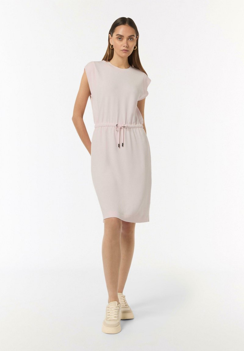 Helles rosa, knielanges Kleid mit kurzen Ärmeln, Bindeband in der Taille und glattem Stoff. Kombiniert mit beigen Schnürsneakern.