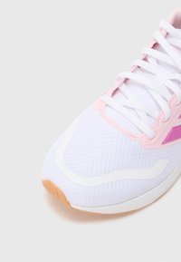 Chaussure de sport blanche avec des accents roses, tige en mesh texturé, lacets plats et semelle extérieure en caoutchouc. Présente des éléments de design blancs incurvés.