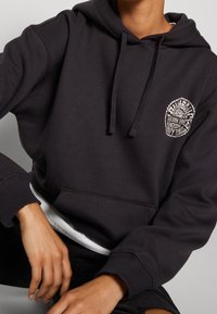 Osoba oblečená v černé mikině s kapucí, na hrudi má bílé kruhové logo a pod tím je vidět bílá košile, ruce má položené na kolenou.