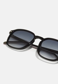 Occhiali da sole oversize neri con lenti sfumate, una robusta montatura in plastica e sottili accenti in metallo sulle tempie. Dettagli interni testurizzati.