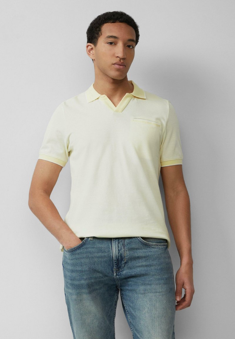 Młody mężczyzna ubrany w jasnoniebieską koszulkę polo z krótkim rękawem i niebieskie dżinsy, z jedną ręką w kieszeni, stojący na tle jasnej, gładkiej ściany.