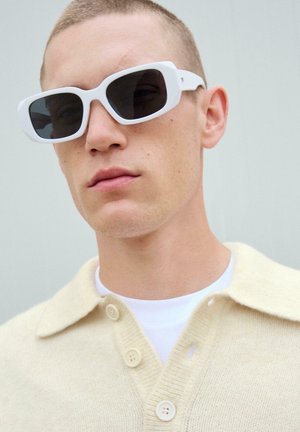 Witte zonnebril met donkere glazen, gedragen met een crèmekleurige gebreide cardigan met een puntkraag en knoopsluiting. Glad huidoppervlak.