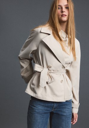 Jeune femme aux longs cheveux blonds portant une veste beige ceinturée et un jean bleu sur un fond gris.