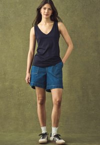 Mørkeblå ærmeløs top med V-udskæring, parret med blå denimshorts med frontlommer. Modellen har hvide ankelstrømper og casual sneakers på.
