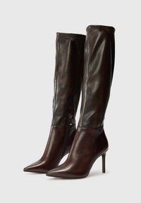 Botas de cuero marrón hasta la rodilla con puntera afilada, tacón de aguja y detalle de cremallera lateral. Textura suave y diseño elegante.