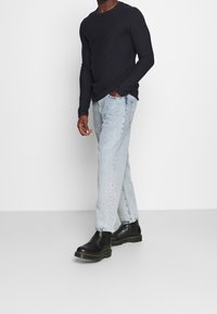 Haut à manches longues bleu marine, jean droit bleu clair, et bottines en cuir noir à semelles épaisses, vus en angle alors que la personne marche.