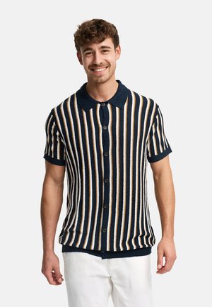 Smilende mand iført en kortærmet, knappet skjorte med lodrette navy-, hvide og beige striber, kombineret med hvide bukser.