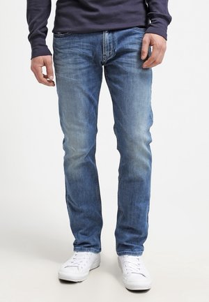 Mann trägt ausgewaschene blaue Jeans, weiße Sneakers und ein marineblaues Langarmshirt, steht vor einem schlichten weißen Hintergrund.