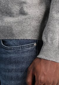 Maglione grigio di lana con polsini a coste, indossato sopra jeans in denim blu con un bottone in metallo e cuciture a contrasto lungo la tasca.