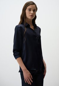 Donkerblauwe satijnen shirt met een klassieke kraag, knopenfront en opgerolde mouwen. Glad textuur met een getailleerde pasvorm.