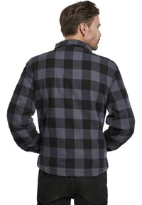 Camisa de franela negra y gris, con un patrón de cuadros, mangas largas y cuello. La parte de atrás muestra un ajuste relajado y un dobladillo curvado.