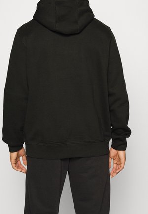 Personne vue de dos portant un sweat à capuche noir et un pantalon noir, les mains détendues le long du corps, sur un fond uni.