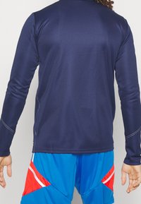 Marinblå långärmad sporttröja med plana sömmar. Har fukttransporterande tyg. Kombinerad med blå shorts med röda och vita detaljer.