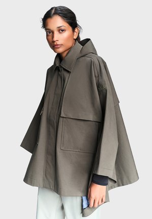 Žena nosi voľný olivovo zelený plášť s kapucňou a veľkými vreckami vpredu a svetlé nohavice, stojaca pred jednoduchým pozadím.