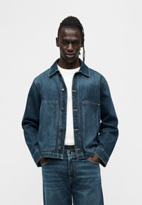 Σκούρο μπλε τζιν μπουφάν με γιακά, κουμπιά μπροστά και δύο τσέπες στο στήθος, συνδυασμένο με λευκό πουκάμισο, πάνω σε μονόχρωμο φόντο.