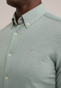 Man i en ljusgrön, knappar polo-tröja med knäppbar krage och broderad fågel-logotyp på bröstet.