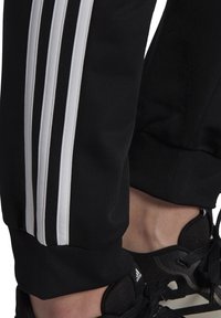 Černé sportovní kalhoty z hladkého polyesteru mají po straně tři bílé pruhy, elastický lem u kotníku a jsou kombinovány s černými teniskami.