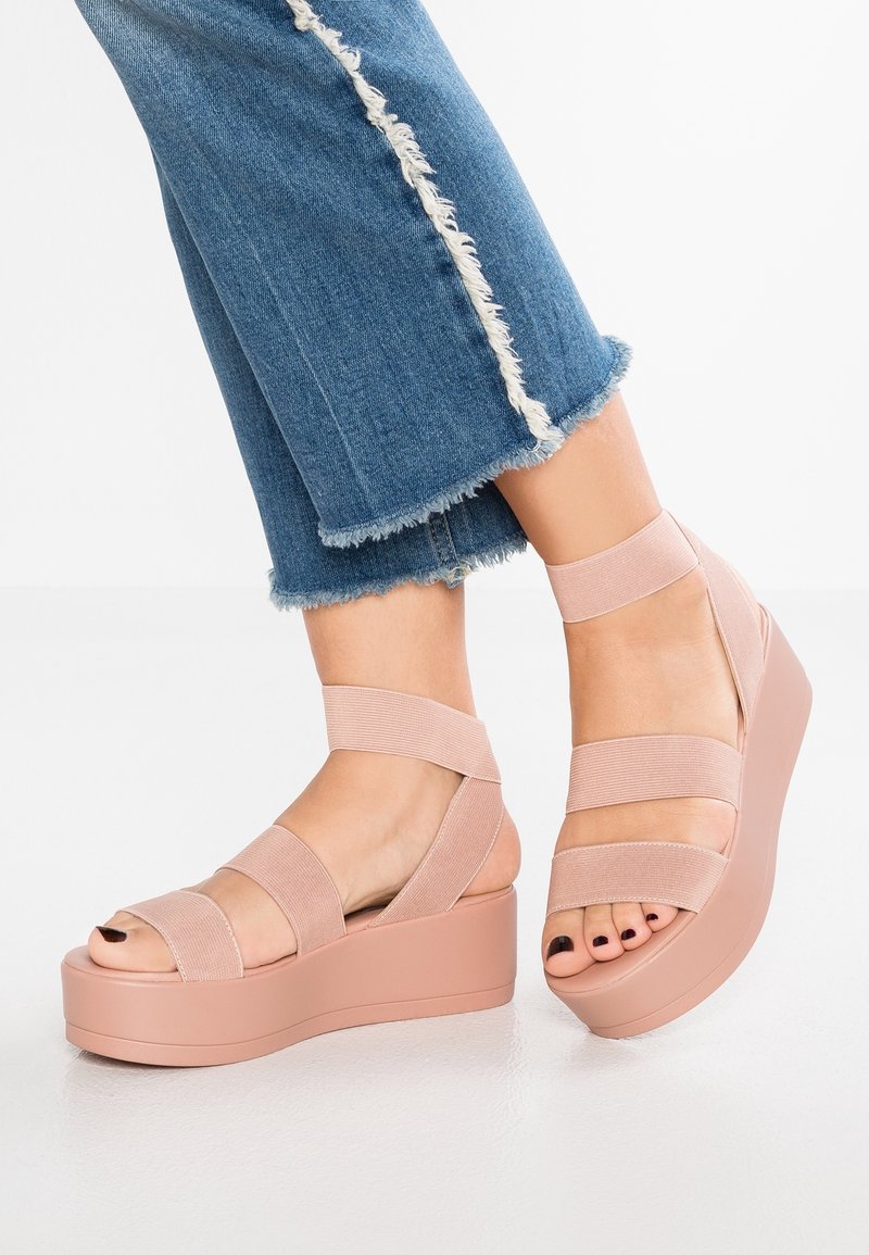 Sandales à plateforme roses avec larges sangles élastiques et finition mate lisse. Bout arrondi et talon épais, assorties à un jean bleu à ourlet effiloché.