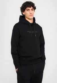 Schwarzer Hoodie aus weichem Stoff mit Kängurutasche und Kapuze mit Zugband. Mit eingeprägtem Markenlogo auf der Brust.
