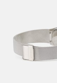 Srebrny meshowy pasek do zegarka o gładkim, teksturowanym wzorze. Wyposażony w metalowy zamek i wygrawerowaną nazwę marki. Gładkie, polerowane akcenty metalowe.