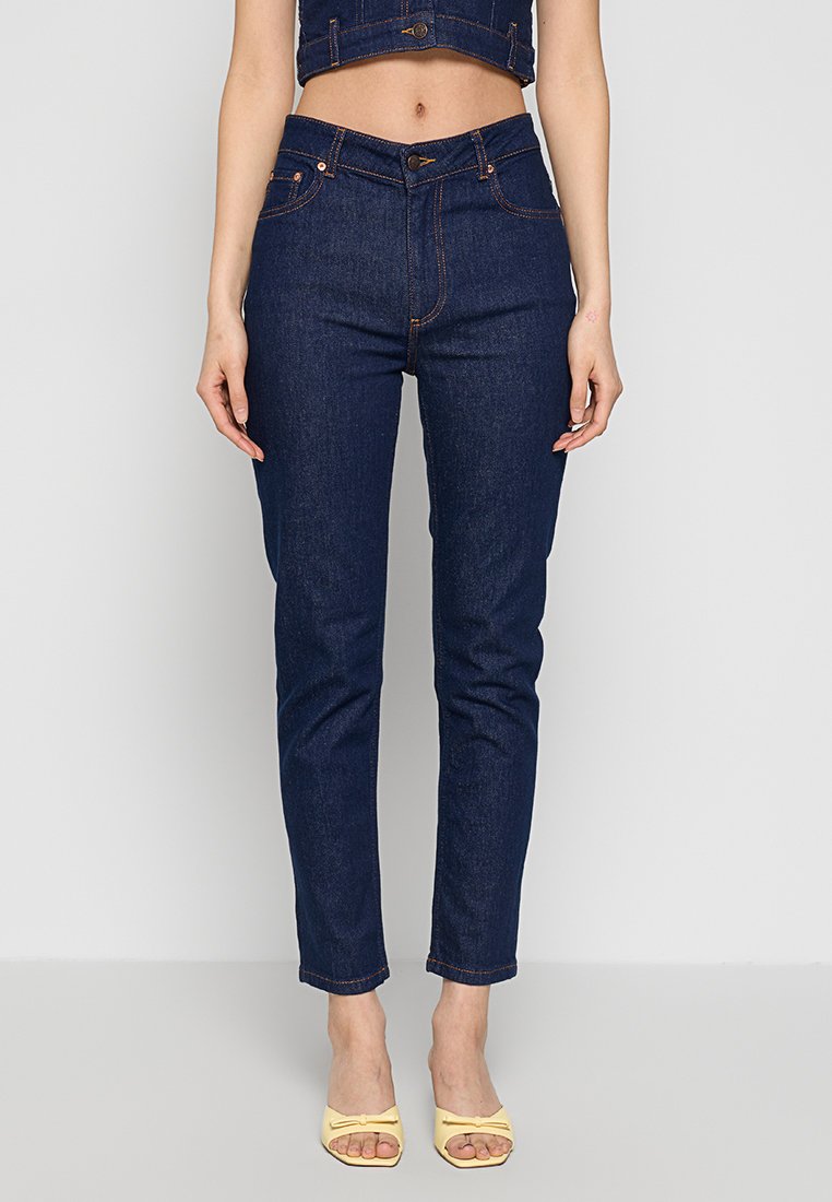 Kobieta w ciemnoniebieskich dżinsach z wysokim stanem i żółtych sandałach na obcasie na tle jasnoszarym.