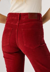 Dettaglio ravvicinato di una persona che indossa pantaloni in velluto a coste rossi a vita alta, con tasche posteriori e un'etichetta di marca in pelle marrone sulla vita.