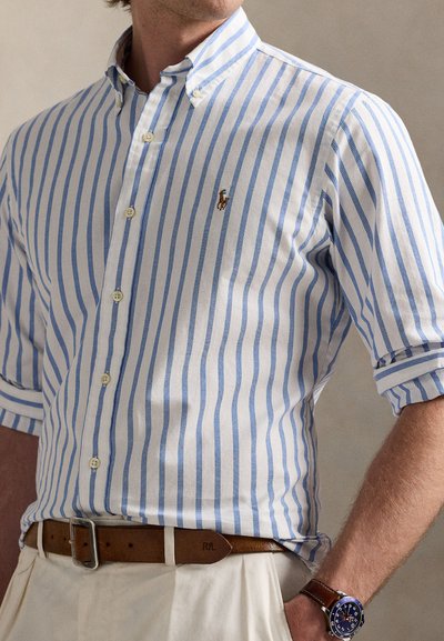 Proužkovaná košile s knoflíky v bílé a světle modré barvě, s malým vyšívaným logem na levé straně hrudi a vyhrnutými rukávy.