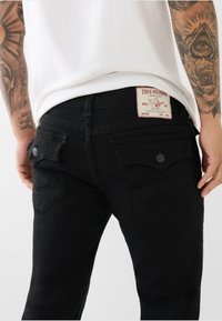 Blugi negri din denim, cu două buzunare pe spate, având un model cusut și o etichetă vizibilă pe talie.