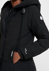 Svart puffjacka med huva, utrustad med en quiltad design, ribbade muddar och en logotyp-patch på ärmen. Dragkedja och tryckknappar.