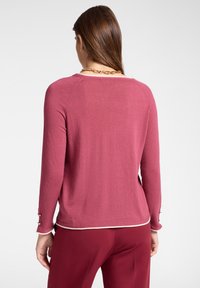 Maglione a maniche lunghe di colore rosa con scollo arrotondato, dettagli decorativi sui polsini e una finitura bianca a contrasto sul bordo.