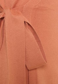 Rustfärgad stickad plagg med en knyten rosett vid midjan, med mjuk textur och en slät yta, accentuerad av ett smalt, loopat band.