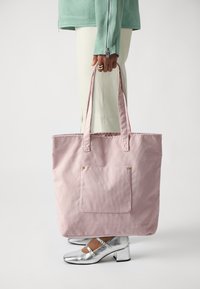 Sac en toile en velours côtelé rose avec deux poignées, comportant une poche avant et des accents en matériel doré, sur un fond neutre.