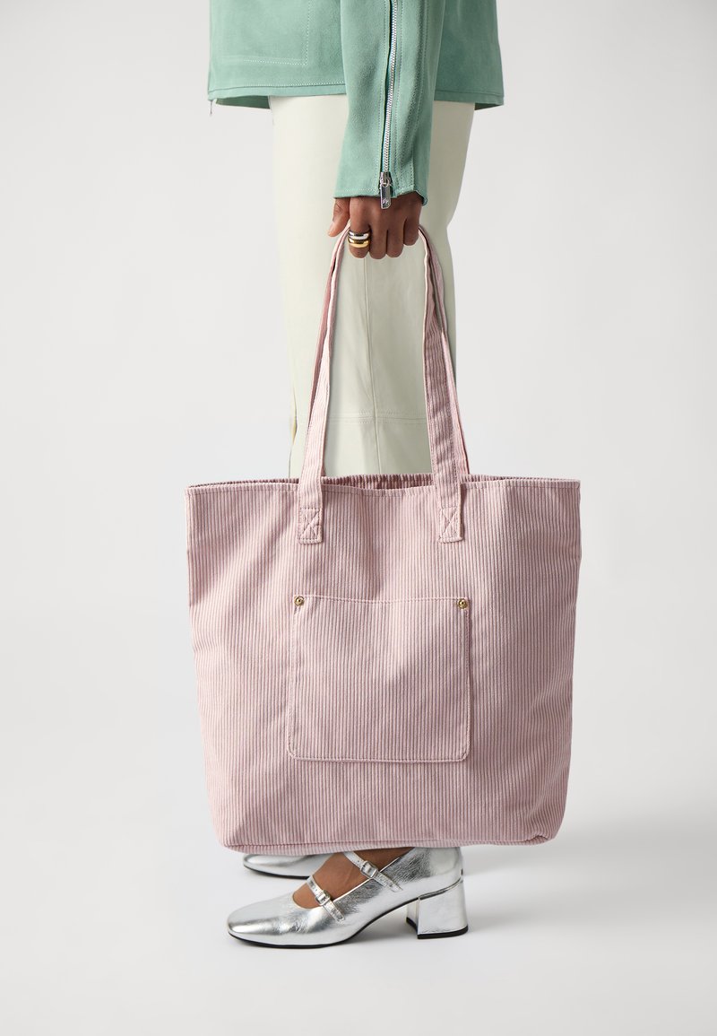 Sac en toile en velours côtelé rose avec deux poignées, comportant une poche avant et des accents en matériel doré, sur un fond neutre.