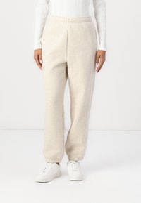 Beige Jogginghosen mit elastischem Bund und Bündchen, aus weichem, strukturiertem Stoff; getragen mit weißen Sneakers vor einem einfarbigen Hintergrund.